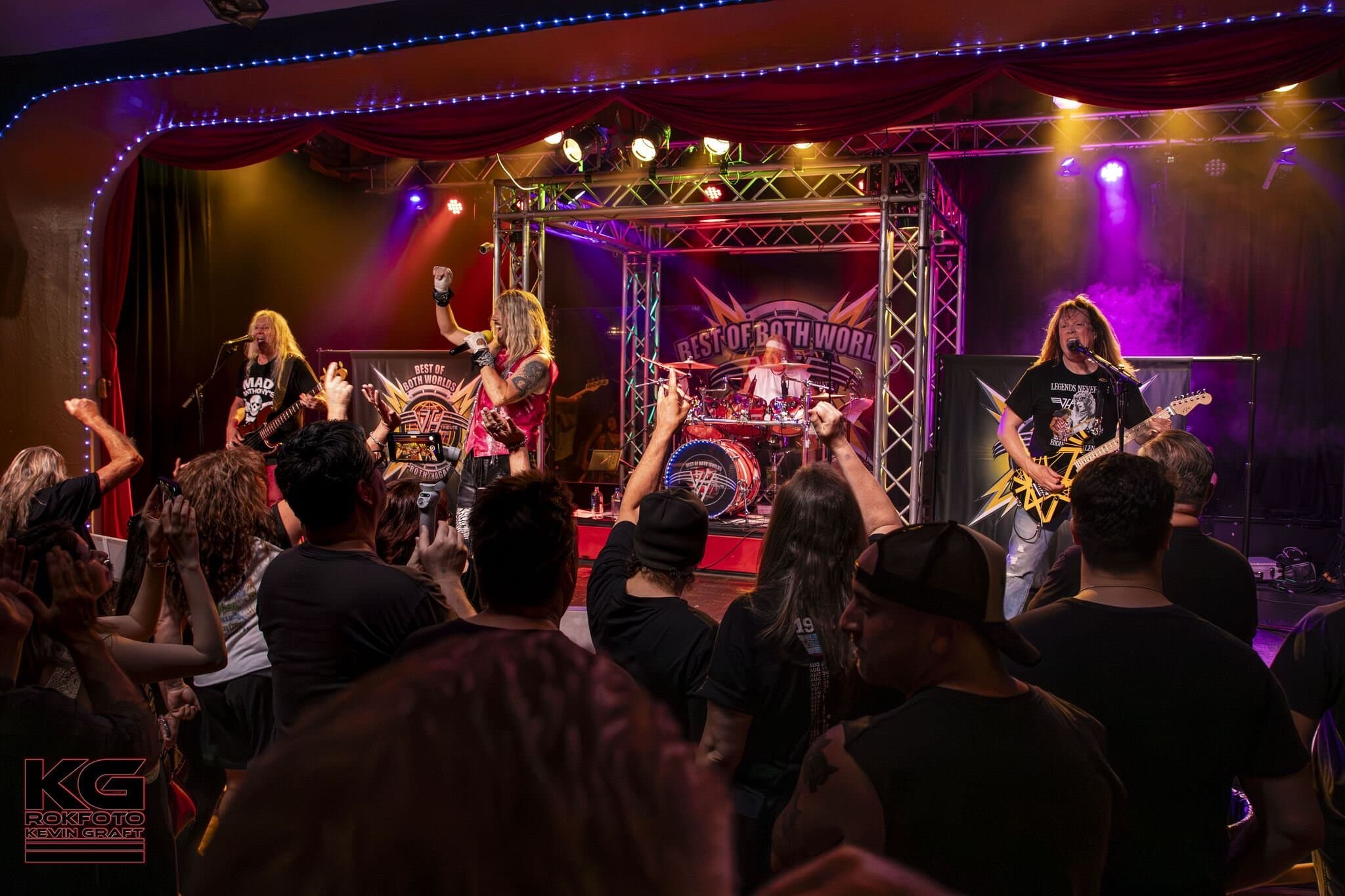 Best of Both Worlds performing live — audience response with hands raised, all four band members on stage with branded kick drum visible